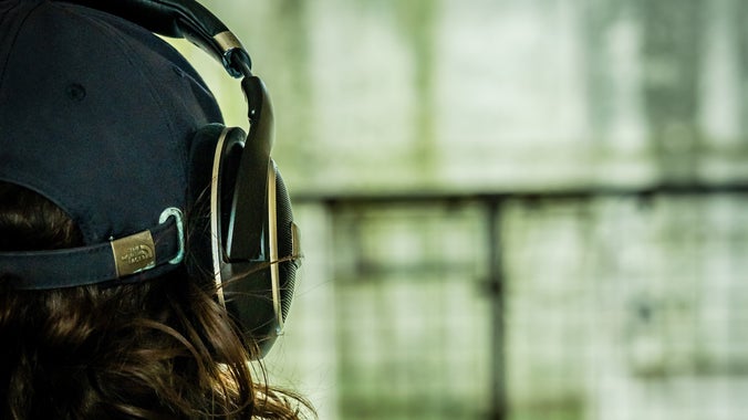 A dark haired woman wearing a baseball cap with large headphones on over the top of her head while listening to an audio play in front of Lab 1, a former military testing laboratory on Orford Ness